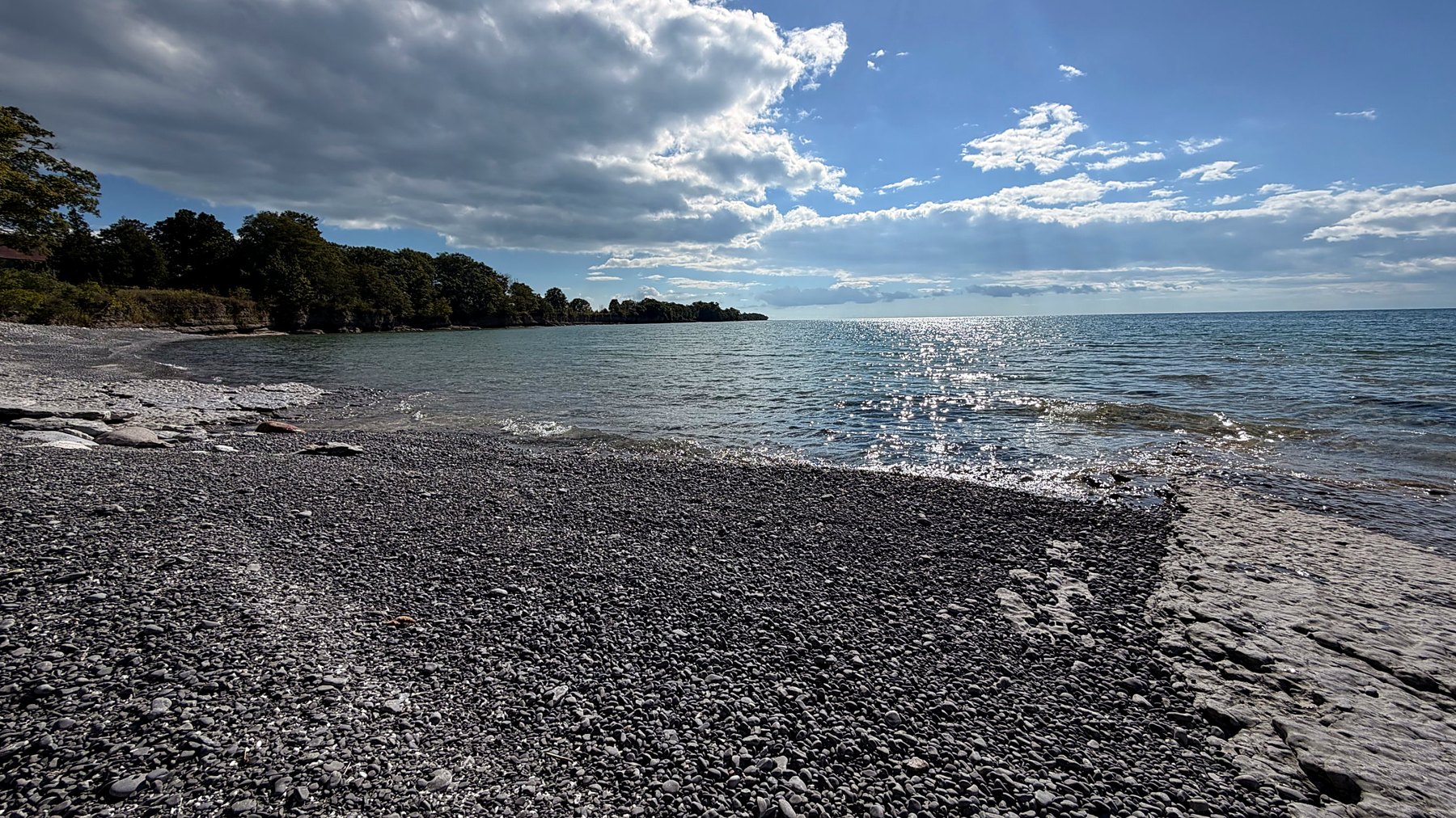 A cobble shoreline at low water, calm lake beyond under broken cloud